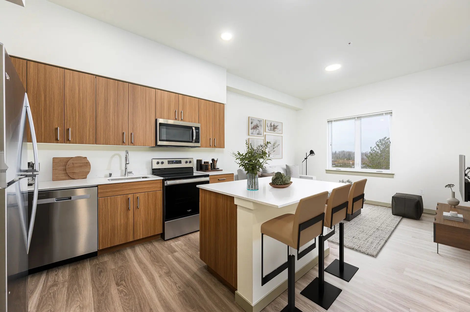 Open kitchen with brown cabinets, wood style flooring, a large island with white countertops and three stools at the bar. The living room is adjacent to the right.