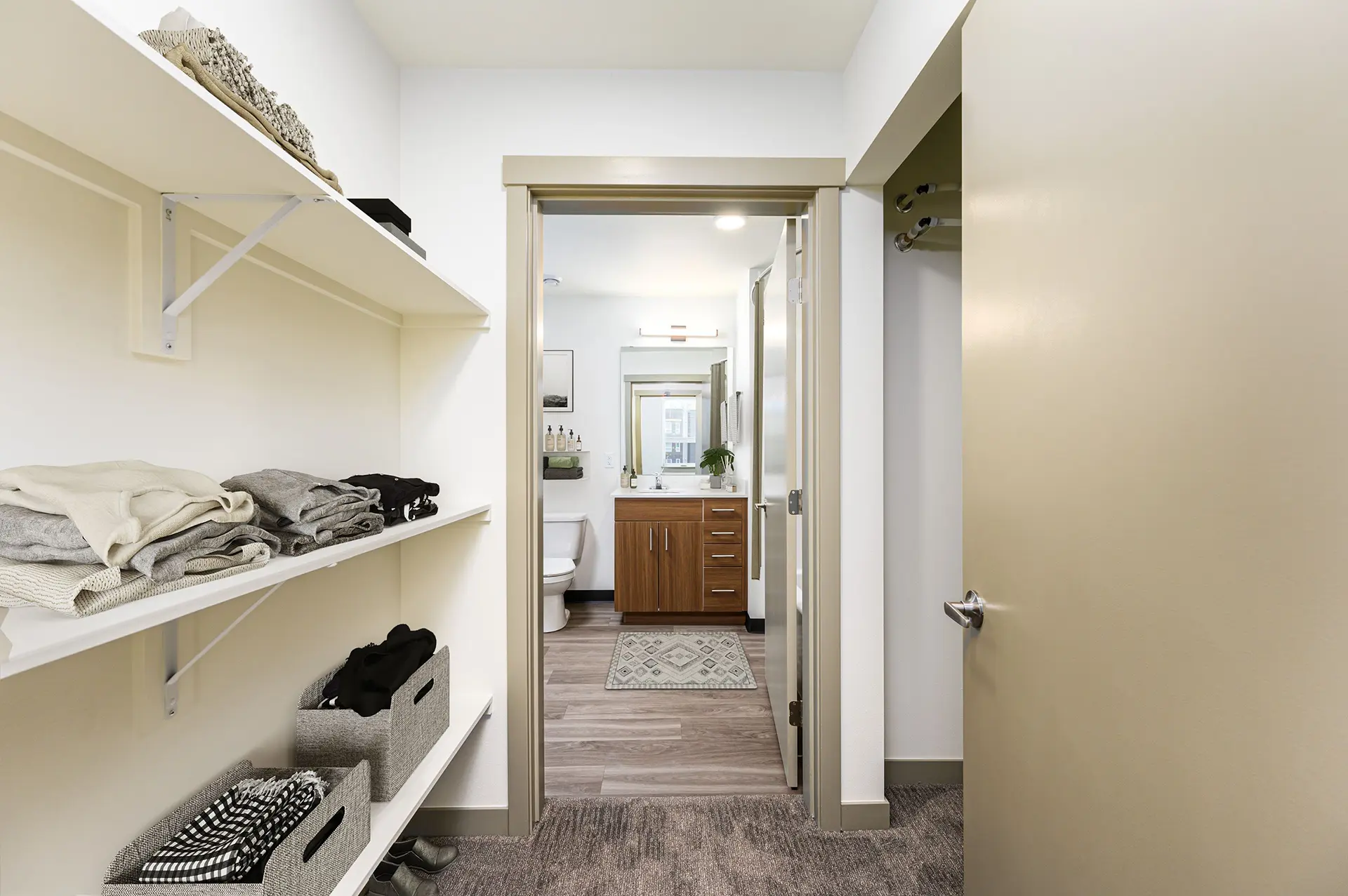 three long shelves leading down a wall to a bathroom with toilet next to single vanity, and an open closet on the right.