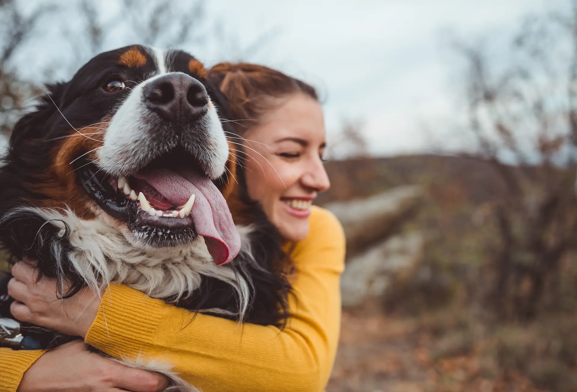 A woman in a mustard yellow sweater outside hugs her large dog with white muzzle and brown and black fur around the eyes.