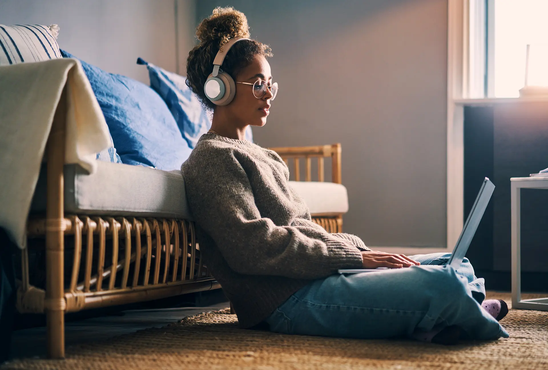 A black woman wearing over ear headphones sits on the floor resting against her couch while working on a notebook computer in her lap.