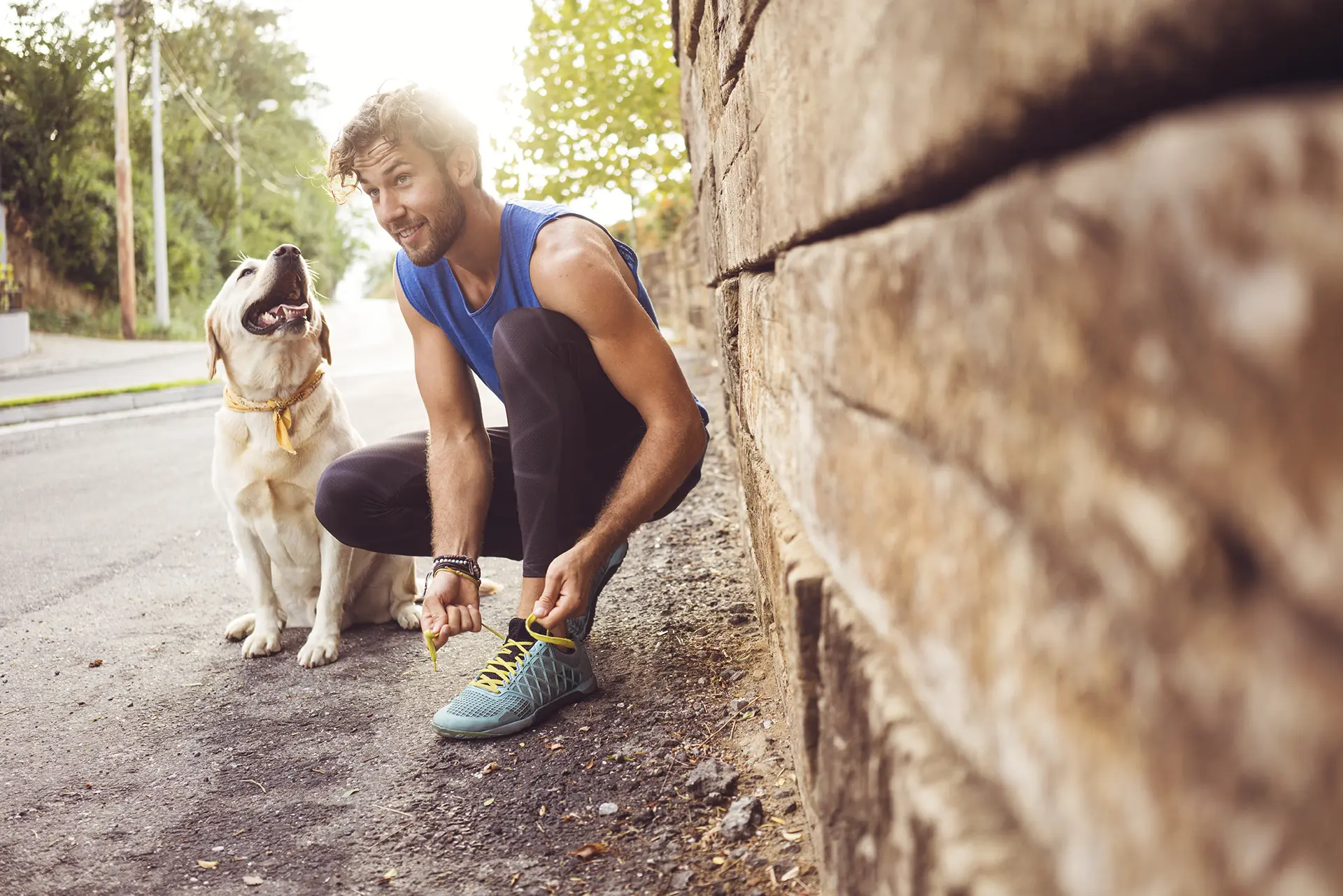 A young man with a short beard wearing fitness attire squats next to his yellow dog and ties his shoe.