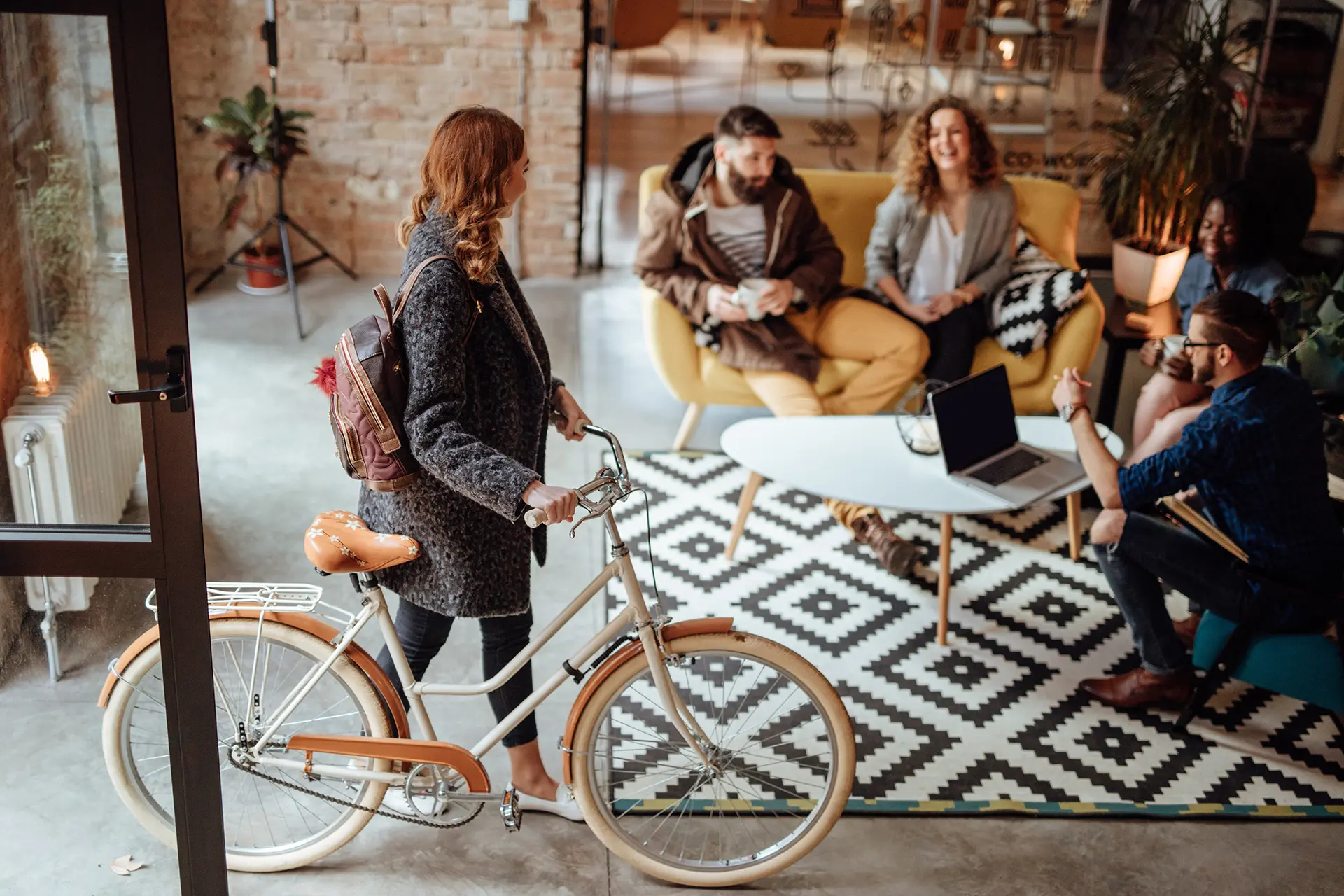 A woman walks her peach and cream cruiser bicycle into a lobby with cement floors and a group of people sitting around a coffee table with a computer.