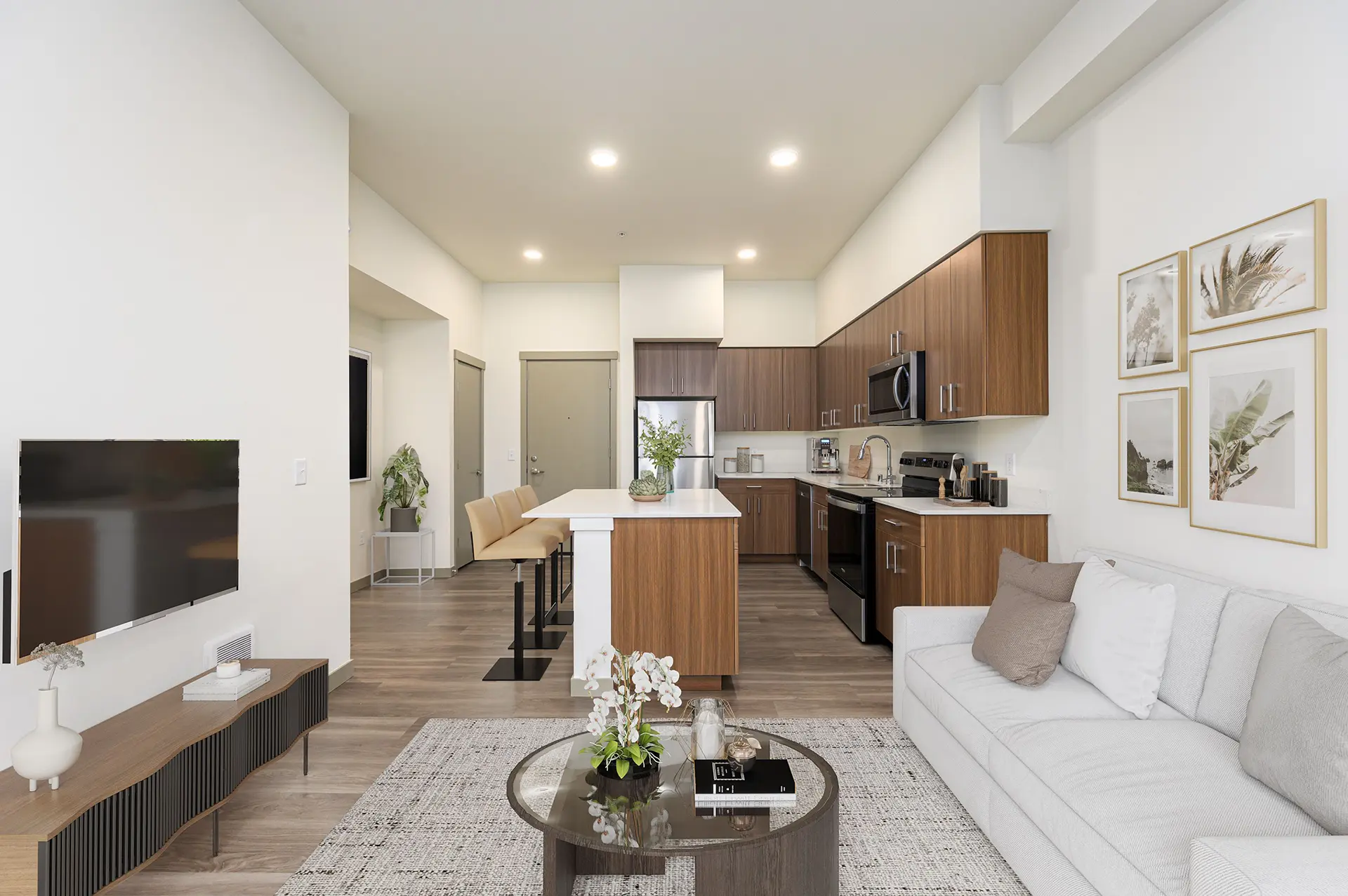 living room with large couch, coffee table, and wall mounted TV. In the background you can see the open kitchen with bar seating at a large island