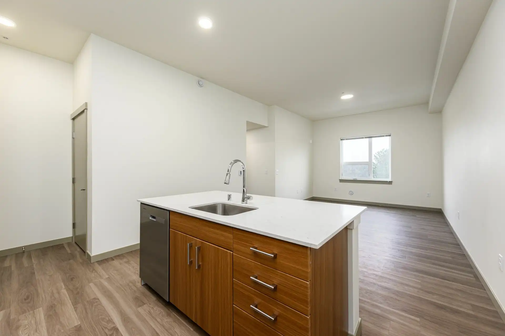 kitchen island with white countertop and brown wood grain custom cabinets, and living room with window in the background. The room has wood style flooring throughout.