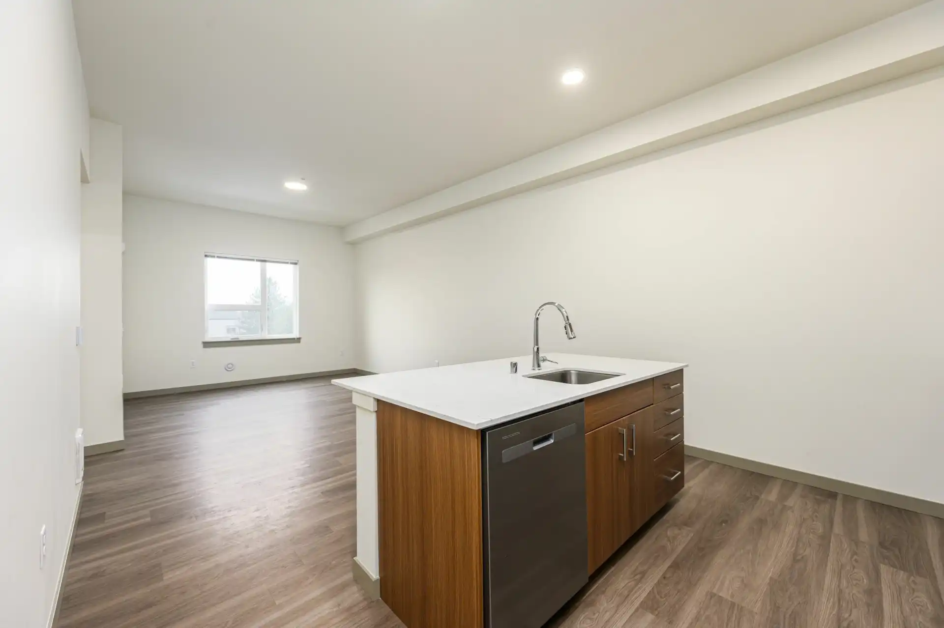 kitchen island in an open room with white countertop, sink, stainless dishwasher, and custom brown wood grain cabinets with brushed nickel hardware.