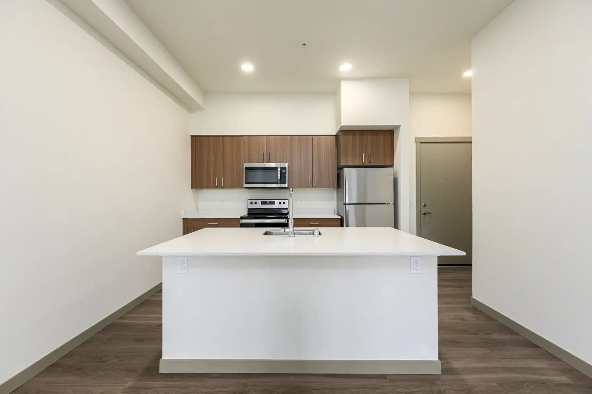 kitchen island with sink in a large white countertop, custom brown cabinets, stove, microwave, and refrigerator line the wall behind with front door to the right