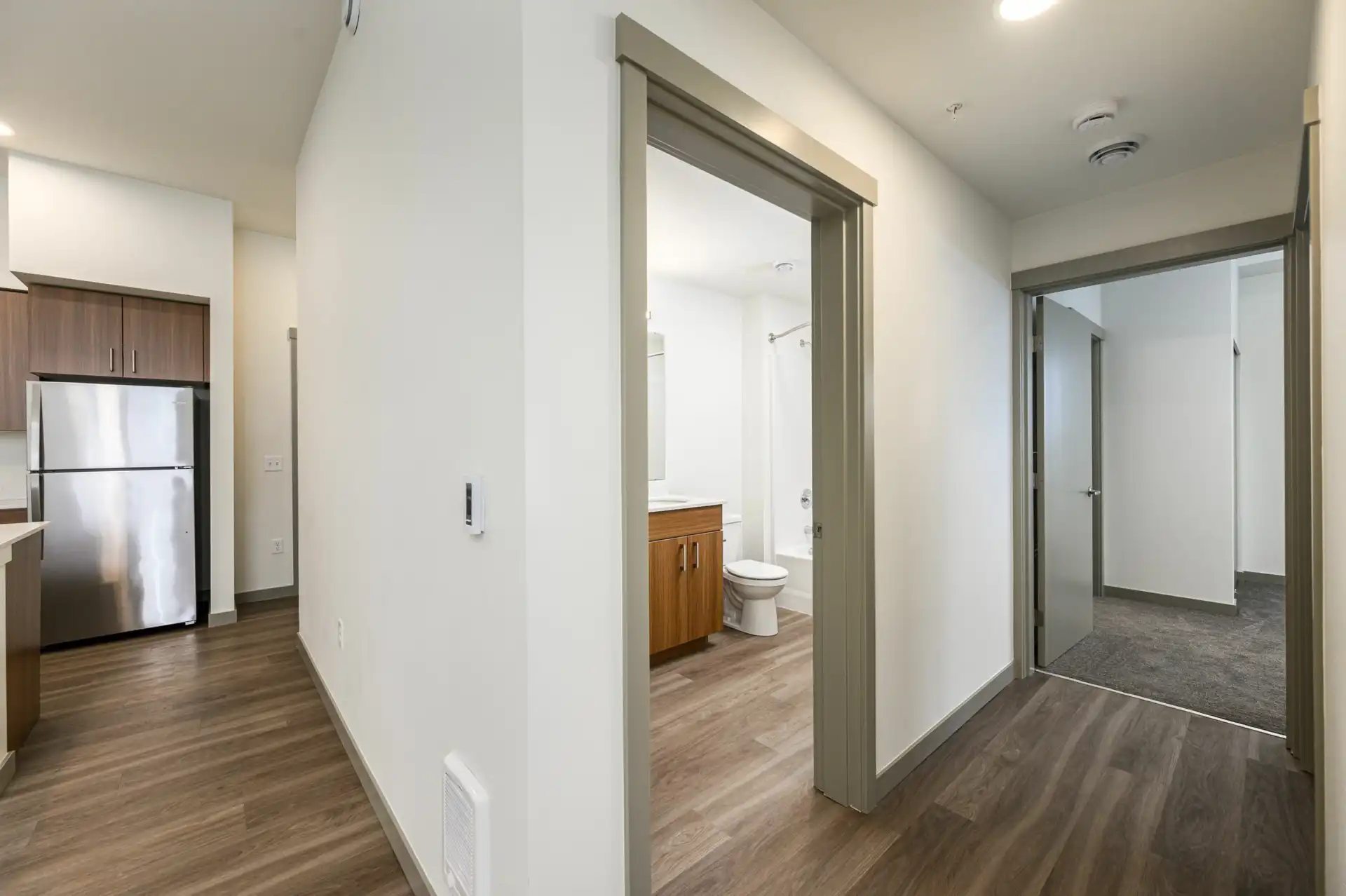 corner view with open kitchen on the left, hallway bathroom in the middle, and open door to a bedroom on the right down the hallway.