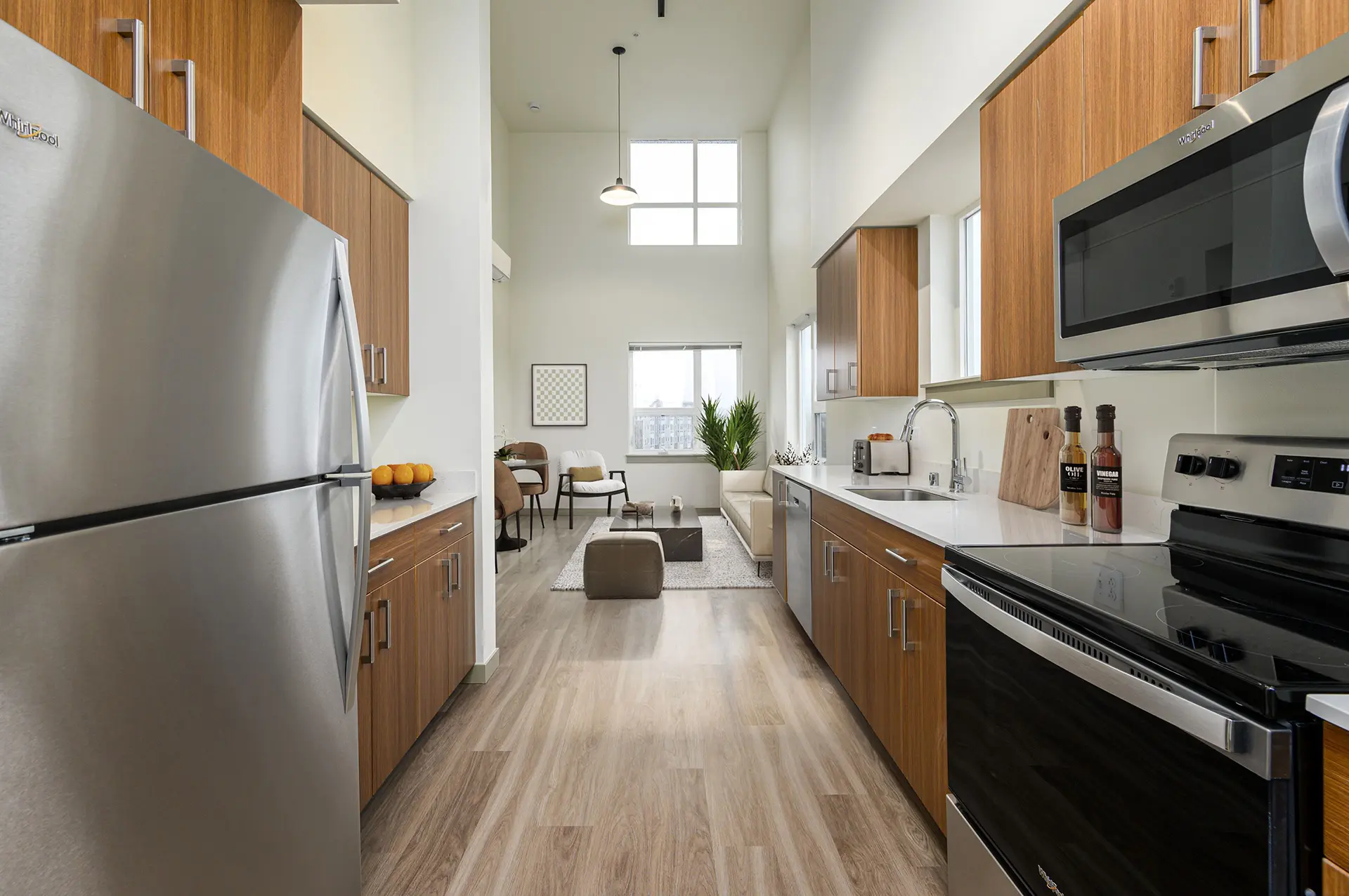 large galley kitchen opening to the living room with custom light brown cabinets, soaring ceiling, white countertops, tan wood flooring, and stainless appliances with glasstop stove.