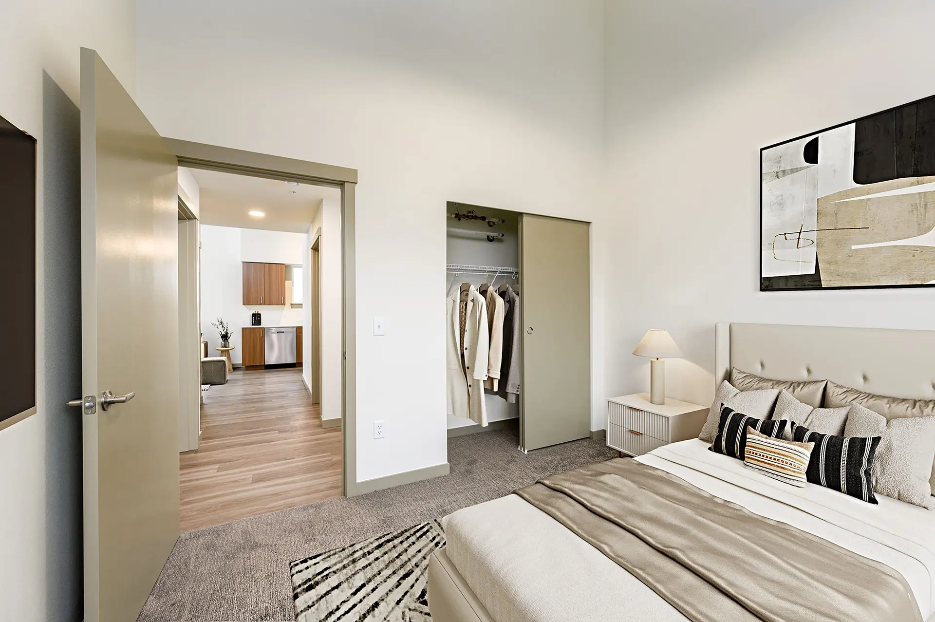 gray carpeted bedroom with sliding door closet, queen sized bed, cream colored nightstand, and abstract painting hanging above the bed.