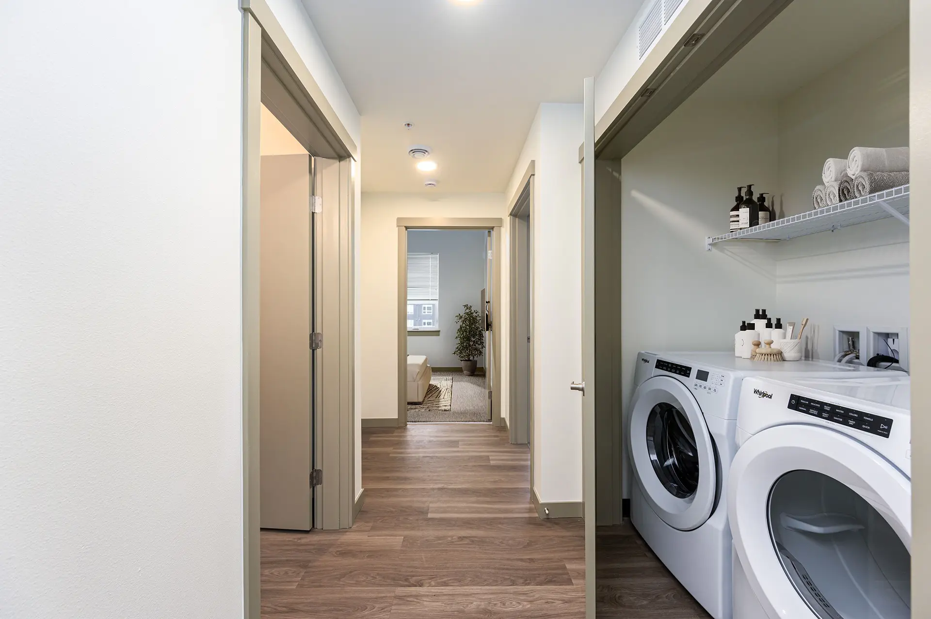 wood floor hallway to bedroom with double-door utility closet containing full size from loading washer and dryer under a wire shelf.