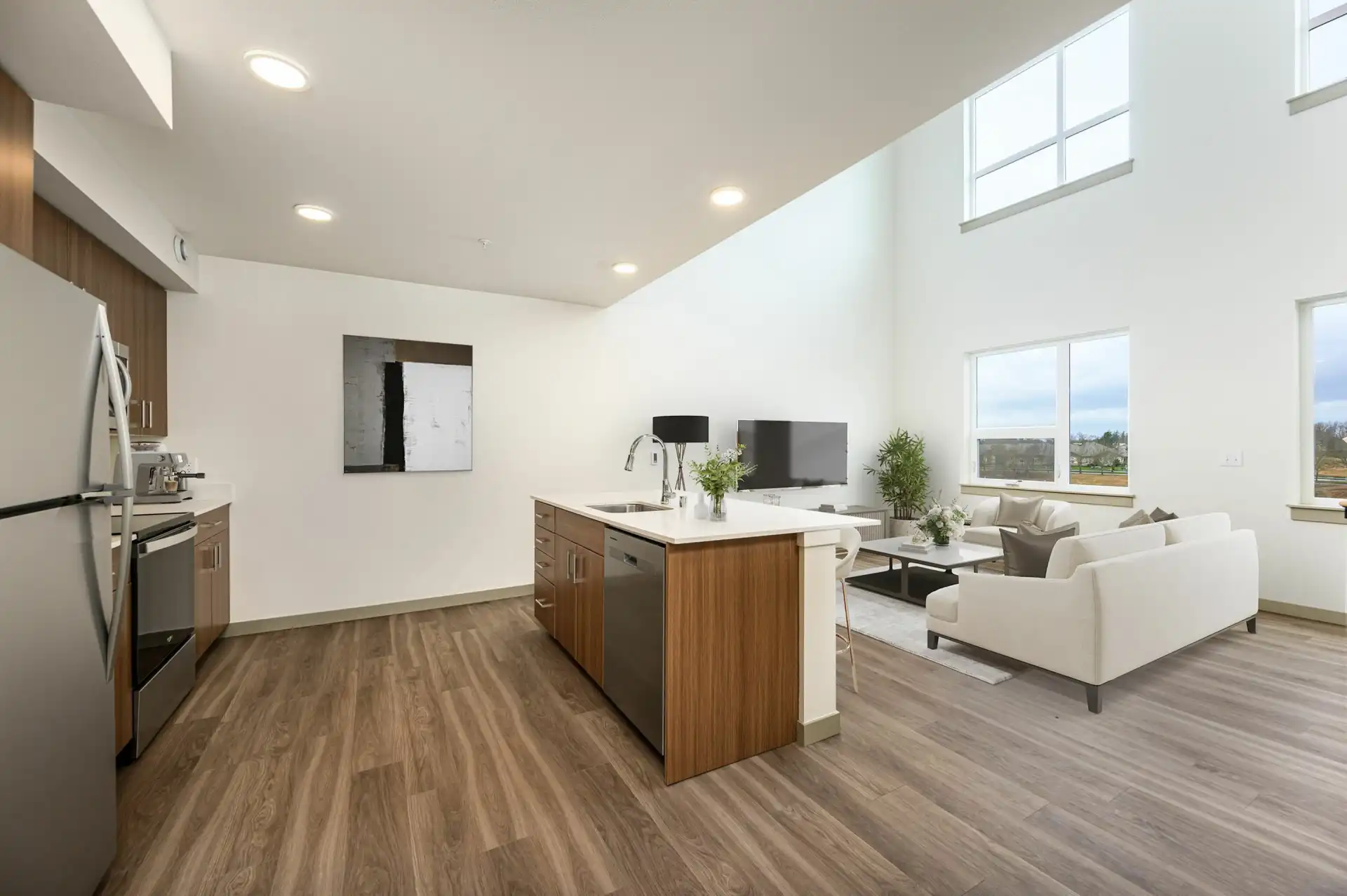 lower level of a loft apartment with open kitchen with large island separating it from the living room. The living room has a high ceiling and four big windows stacked in rows of two.