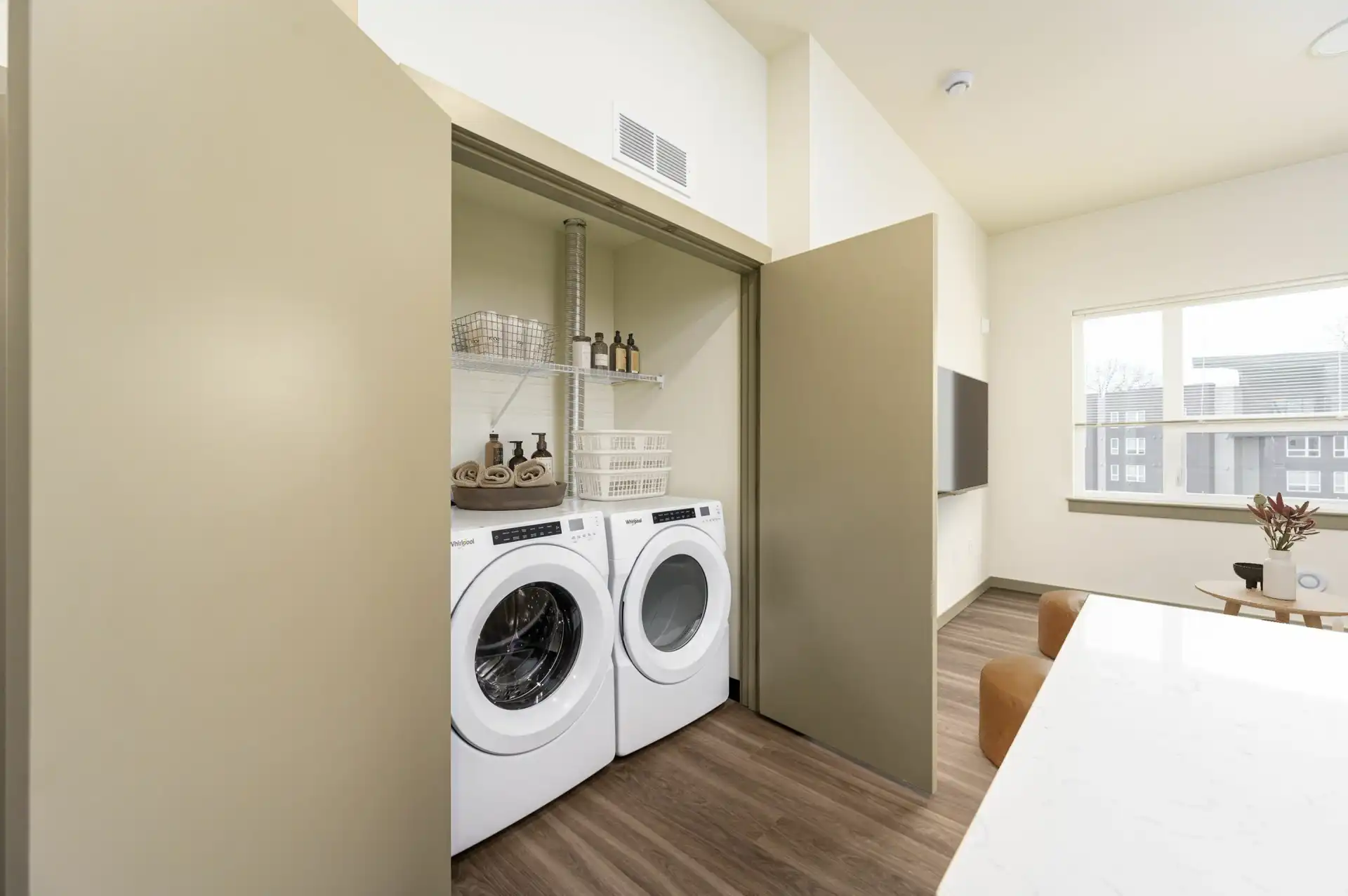 double door utility closet next to a kitchen, with full size front loading washer and dryer in the closet under a wire shelf. The living room with wall mounted TV is in the background.